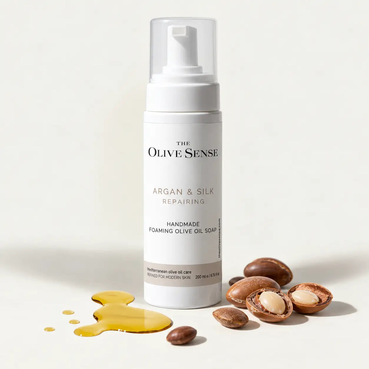 The Olive Sense Argan & Silk Repairing Handmade Foaming Olive Oil Soap in a white pump bottle, styled with argan nuts and a pool of golden olive oil on a clean white background.