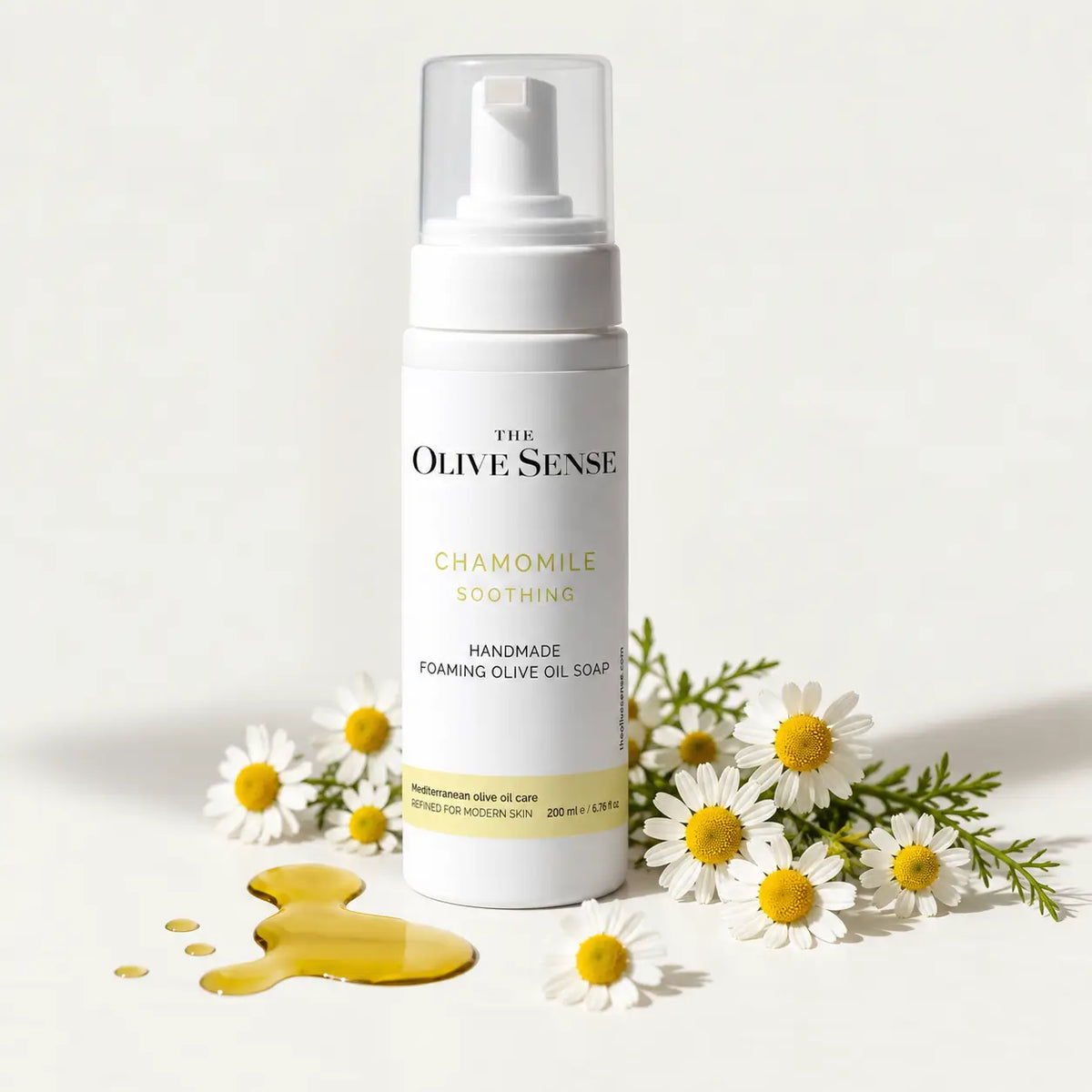 A bottle of The Olive Sense Chamomile Soothing Handmade Foaming Olive Oil Soap stands on a white surface with chamomile flowers and a pool of golden olive oil around it.