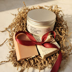Jar of body cream, heart-shaped soap, and square soap with a red ribbon on a bed of straw.