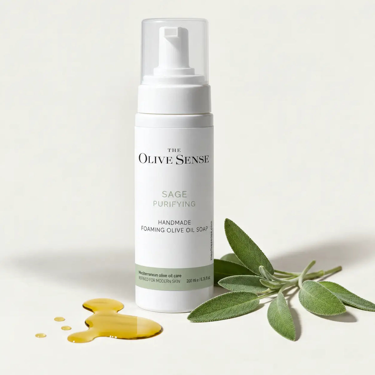 A white pump bottle of The Olive Sense Sage Purifying Handmade Foaming Olive Oil Soap, styled with a small pool of golden olive oil and fresh sage leaves on a clean, off-white background.