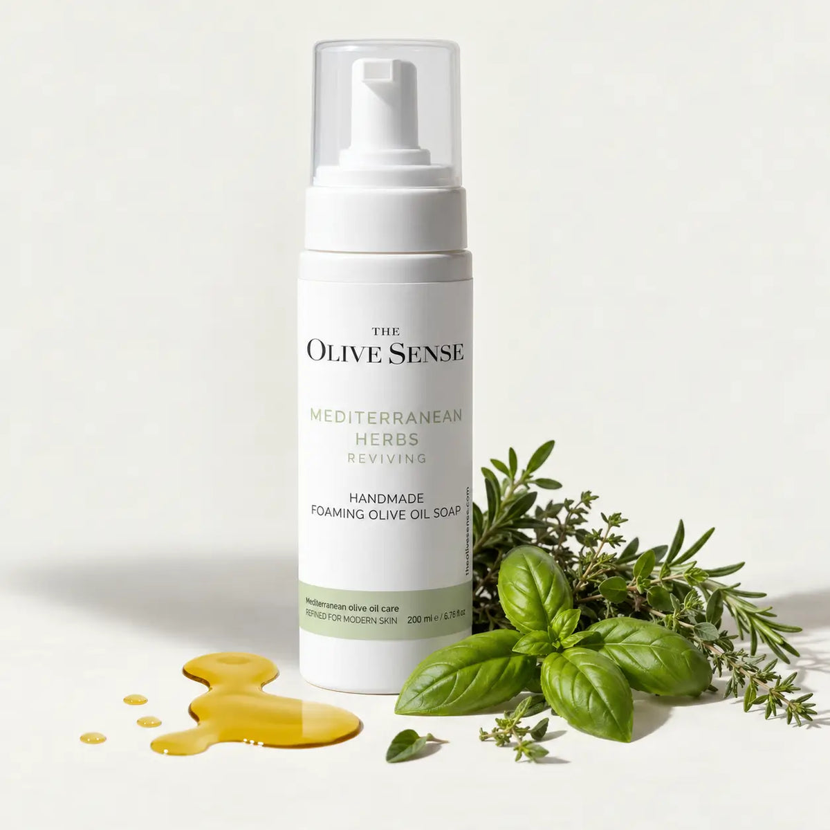 A bottle of The Olive Sense Mediterranean Herbs Reviving Handmade Foaming Olive Oil Soap stands on a white surface with olive oil droplets and fresh green herbs like basil and thyme arranged around it.